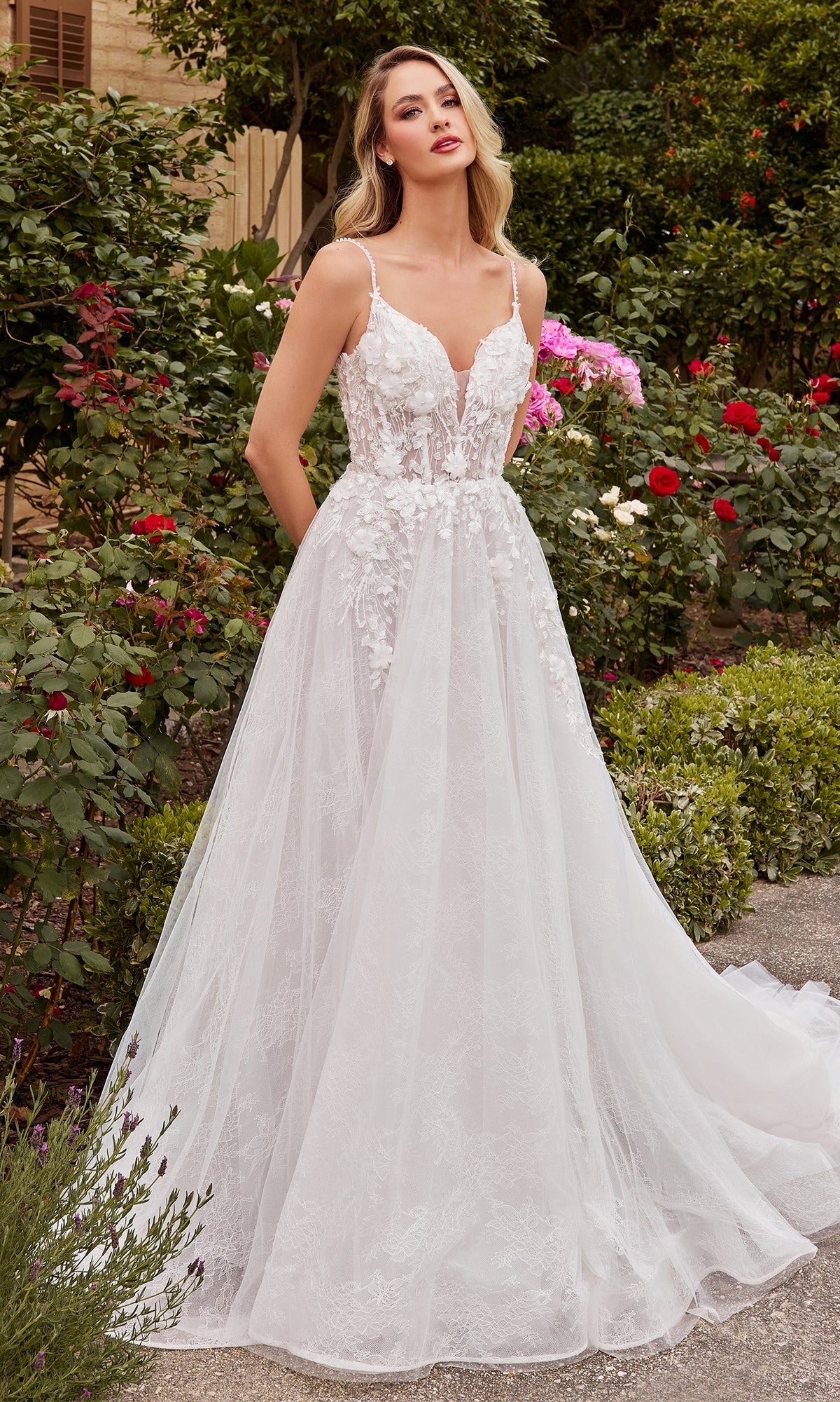 A woman stands outdoors in a garden wearing the A-Line White Lace Wedding Gown CD857W, featuring thin straps, 3D floral details, and a sheer corset bodice for an elegant bridal look.
